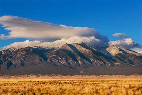 Wide landscape view of a mountain range with rugged peaks under dramatic layered clouds, rising above a dry grassland plain beneath a clear blue sky.