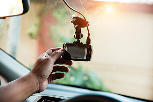 Hand adjusting a small dashboard camera mounted to the inside of a car windshield with a suction cup, the device hanging from a power cable while sunlight filters through the glass and the road outside appears softly blurred.