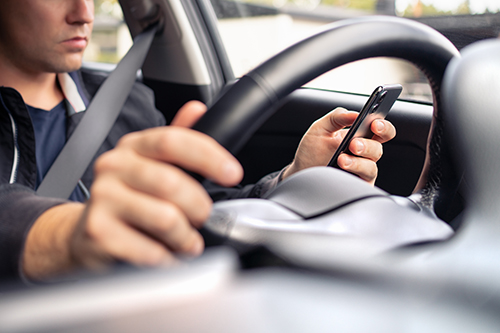 Driver holding a smartphone while steering a car, with one hand on the wheel and eyes focused downward, illustrating distracted driving inside a vehicle.