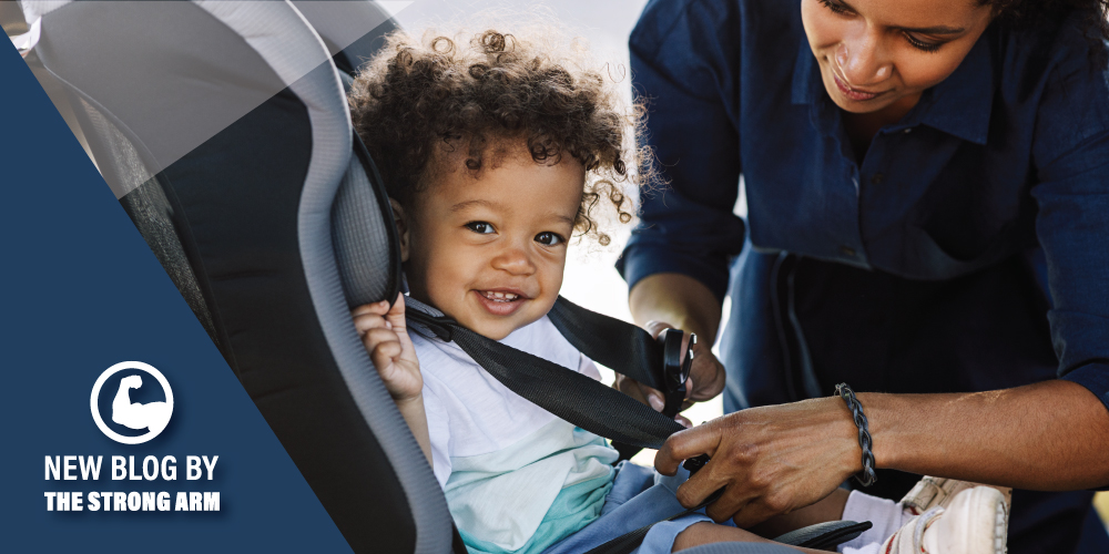 A smiling toddler with curly hair sits secured in a gray child car seat while an adult caregiver leans in to fasten and adjust the black safety harness, with the child looking toward the camera and the adult’s hands visible tightening the straps inside a vehicle interior.