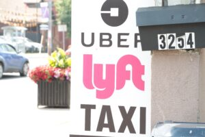 Street-side sign displaying ride-hailing logos for Uber and Lyft above the word “TAXI,” positioned next to a residential mailbox with house number 3254, with a blurred city street, parked car, and flower planter visible in the background.