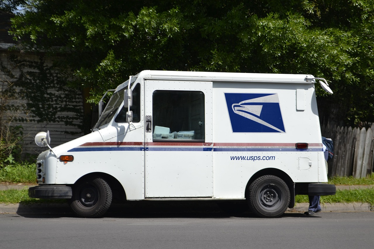 Side view of a white USPS mail delivery truck parked on a residential street, showing the postal service logo and website on the side, with trees and a wooden fence in the background and a postal worker partially visible near the rear of the vehicle.