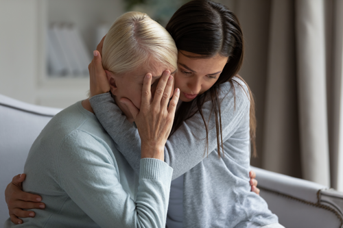 Younger woman comforting an older woman by holding her closely as the older woman covers her face, conveying emotional support and distress in a quiet indoor setting.