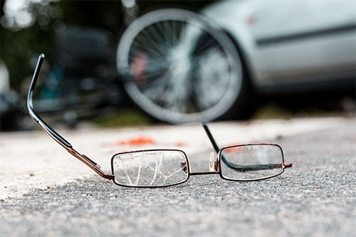 Broken eyeglasses lying on the pavement with a blurred bicycle wheel and vehicle in the background, suggesting the aftermath of a traffic collision involving a cyclist.