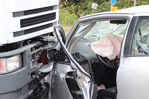A severely damaged white car after a collision with a large truck, showing a crushed front door, shattered windshield, deployed airbag inside the vehicle, and exposed mechanical parts at the point of impact.