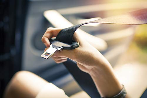 Close-up of a person’s hand pulling a seat belt across their body inside a car, holding the metal buckle near the latch with sunlight streaming through the vehicle interior, emphasizing vehicle safety and restraint use.
