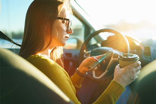 Young woman wearing glasses sits in the driver’s seat of a car holding a smartphone and a takeaway coffee cup while sunlight streams through the windshield.