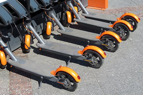 Row of shared electric scooters parked neatly on a sidewalk, showing orange wheel covers, black frames, and folded kickstands in an urban outdoor setting.