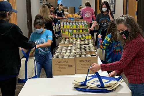 Volunteers wearing face masks pack canned food into reusable bags at long tables inside a community distribution center, with stacked boxes of donated goods in the background.
