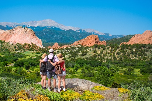 Three hikers stand arm in arm on a rocky overlook, facing a wide landscape of green valleys, red rock formations, and distant mountains under a clear blue sky.