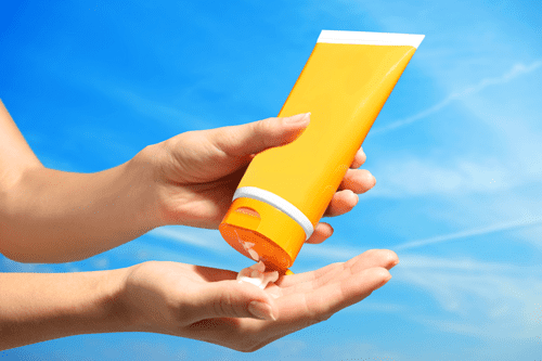 A close-up of hands squeezing white sunscreen lotion from a bright yellow tube into an open palm against a clear blue sky, suggesting sun protection and outdoor skin care.