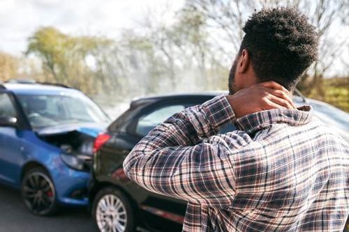Man standing near damaged cars after a collision, holding his neck while looking at the crash scene on a roadway with visible vehicle damage.